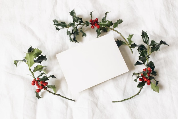 Escena de maqueta de invitación de tarjeta de felicitación festiva con estilo navideño Naturaleza muerta con tarjeta de papel y bayas de acebo sobre fondo de lino blanco Composición de papelería de invierno Vista plana superior