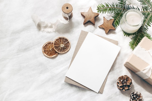 Escena de maqueta de tarjeta de felicitación en blanco de Navidad Composición festiva de boda de invierno Sobre artesanal Caja de regalo de cono de pino Rodajas de fruta naranja y rama de abeto sobre fondo de mantelería blanca Endecha plana