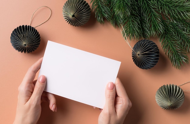 Escena de maqueta de tarjeta de felicitación con manos femeninas sosteniendo una tarjeta de papel en blanco contra adornos navideños