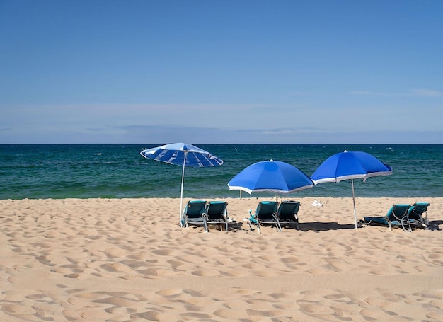 Una escena de playa con una sombrilla azul y sillas en la arena.