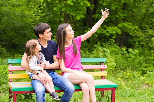 Escena de verano de familia joven feliz tomando autorretratos con su teléfono inteligente en el parque.