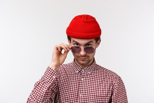 Escéptico y dudoso joven inconformista guapo con bigote, gorro rojo, mira por debajo de la frente con incredulidad, gafas de despegue para hacer una mirada seria de juicio a una persona extraña