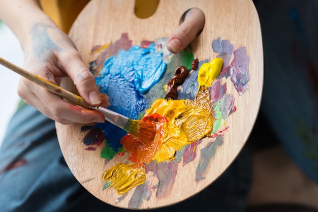 Escola de Belas Artes. Closeup de mãos de artista segurando uma paleta de madeira, misturando tinta acrílica com pincel.