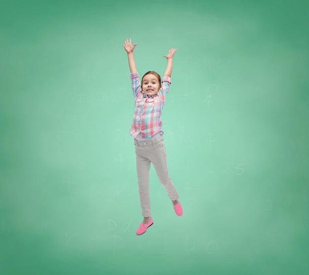 escola, educação, infância, liberdade e conceito de pessoas - menina feliz pulando no ar sobre o fundo verde do quadro de giz escolar