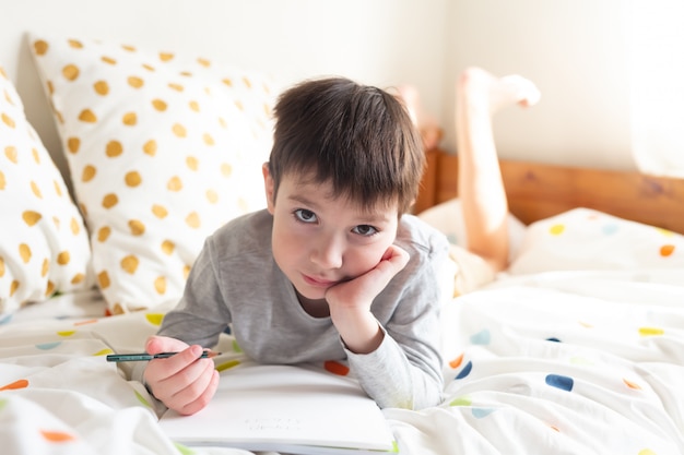 Escolar en la cama y hacer la tarea escolar en casa, escribiendo en el cuaderno. Educación a distancia en línea. Cuarentena