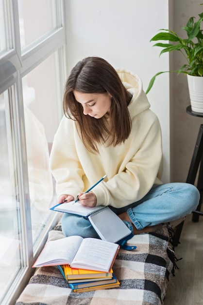 Escolar em casa Uma adolescente estudante senta-se no peitoril da janela com um monte de livros e escreve lição de casa Preparação de ensino à distância para exames Foco seletivo suave