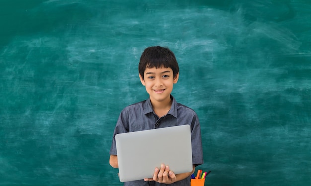 Escolar feliz asiático que sostiene el ordenador portátil en tablero negro