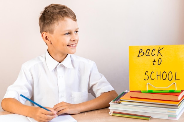 un escolar se sienta en un escritorio contra el fondo de los libros de texto con un cartel amarillo de regreso a la escuela