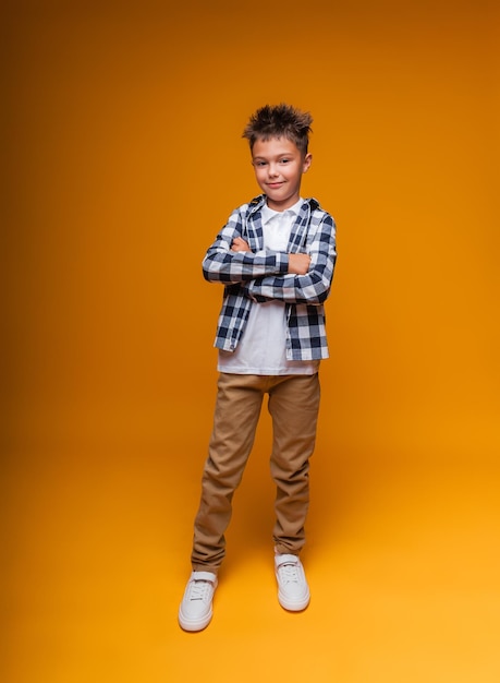 Un escolar sonriente con una camisa a cuadros y una camiseta blanca se para con los brazos cruzados sobre el pecho aislado en un retrato de fondo amarillo de un niño