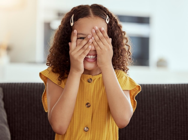 Esconde-esconde retrato de criança e menina com as mãos na cara em uma casa com um sorriso Jogo divertido e criança brincando no sofá da sala com felicidade e diversão em um sofá com a juventude e se escondendo tímido