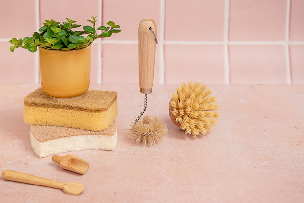 Escovas de limpeza ecológicas naturais de bambu, esponjas, utensílios e uma planta verde em um pote amarelo sobre um fundo de azulejo rosa.