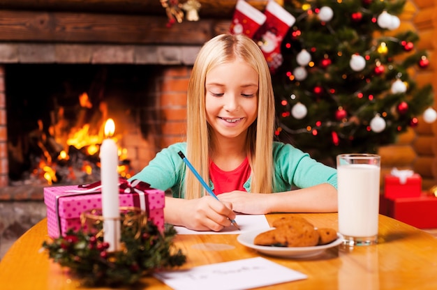 Escrevendo uma carta para o Papai Noel. Menina bonitinha escrevendo uma carta para o Papai Noel enquanto está sentado em casa com a árvore de Natal e a lareira ao fundo