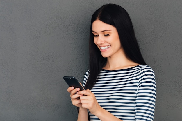 Escribiendo mensaje a un amigo. Atractiva mujer joven sosteniendo un teléfono inteligente y mirándolo con una sonrisa de pie
