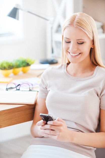 Escribiendo mensaje a un amigo. Hermosa joven sosteniendo un teléfono móvil y sonriendo mientras está sentada en su lugar de trabajo en la oficina