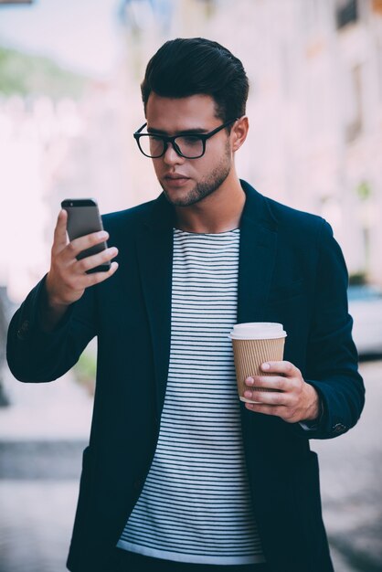 Escribiendo un mensaje rápido a un amigo. Apuesto joven en ropa casual elegante sosteniendo una taza de café y mirando su teléfono inteligente mientras camina por la calle