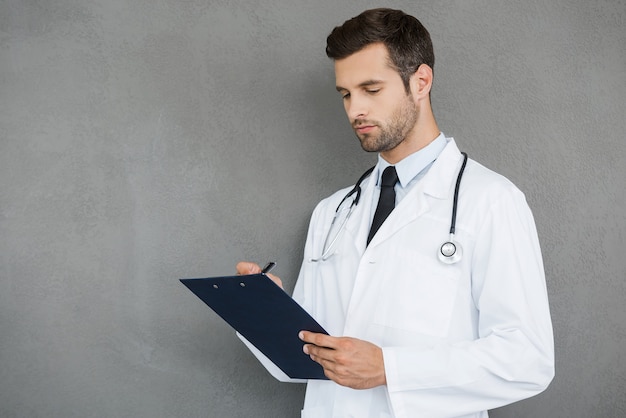 Escribiendo la receta. Seguro médico joven en uniforme blanco escribiendo en el portapapeles mientras está de pie contra el fondo gris