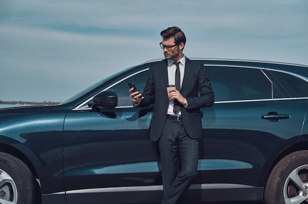 Escribir mensaje comercial. Apuesto joven empresario con teléfono inteligente y tomando café mientras está parado cerca de su coche al aire libre