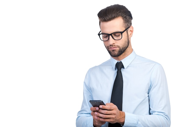 Escribir mensaje comercial. Hombre guapo joven confidente en camisa y corbata sosteniendo el teléfono móvil y mirándolo mientras está de pie contra el fondo blanco