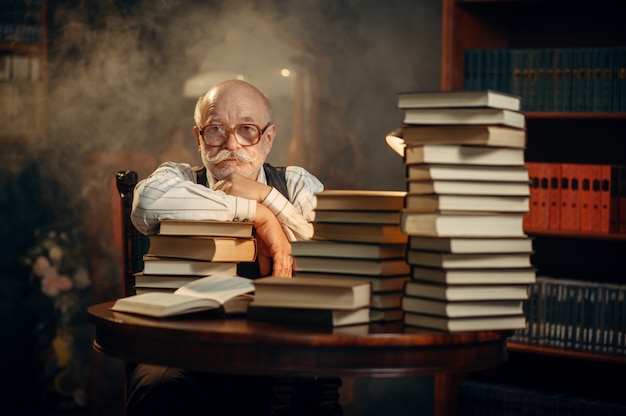 Escritor anciano sentado a la mesa con una pila de libros en la oficina en casa. Anciano con gafas escribe novela literaria en la habitación con humo, inspiración