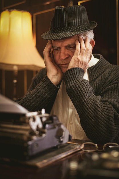 Escritor de homem sênior retrô preocupado com chapéu, sentado à mesa, olhando o texto na máquina de escrever obsoleta e pensando.