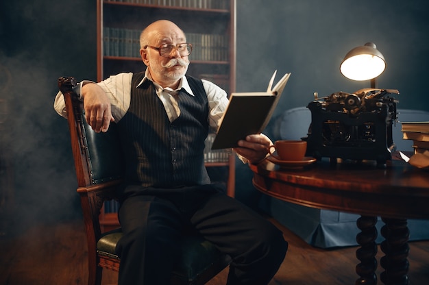 Escritor idoso lê seu trabalho em uma máquina de escrever vintage no escritório em casa. Velho de óculos escrevendo romance de literatura em uma sala com fumaça