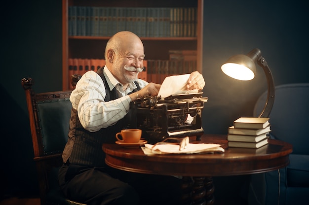 Escritor idoso sorridente trabalha na máquina de escrever retrô no escritório em casa. Velho de óculos escreve romance de literatura em uma sala com fumaça, inspiração