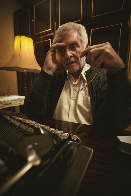 Escritor pensativo Retro Senior Man con gafas en la mano, sentado en el escritorio junto a Obsolete Typewriter y pensando.