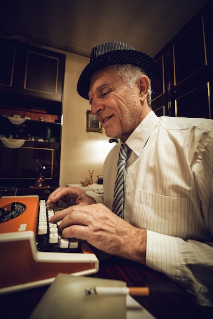 Escritor retro senior con sombrero en su nad, escribiendo en una máquina de escribir obsoleta.