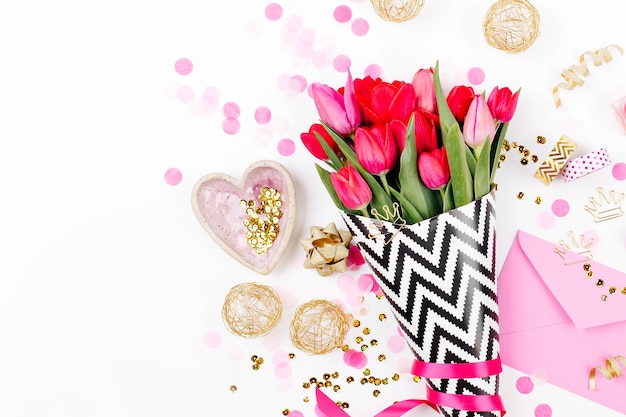 Escritorio de estilo rosa y dorado con flores. Tulipanes rosados en papel de regalo con estilo blanco y negro, regalos, cosméticos y accesorios femeninos con confeti sobre fondo blanco.