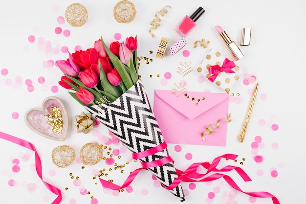 Escritorio de estilo rosa y dorado con flores. Tulipanes rosados en papel de regalo con estilo blanco y negro, regalos, cosméticos y accesorios femeninos con confeti sobre fondo blanco.