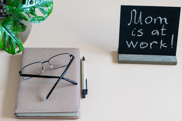 Escritorio, lugar de trabajo a domicilio con diario de tiempo, bolígrafo, gafas y negro en blanco con inscripción Mamá está en el trabajo. Concepto de trabajo remoto en casa.