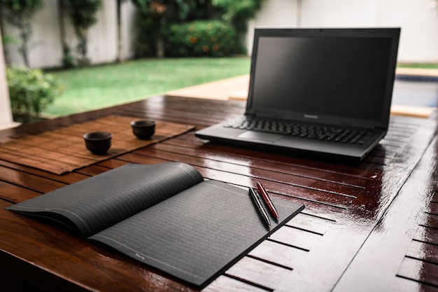 Un escritorio de madera al aire libre con un cuaderno negro, dos bolígrafos y vasos de arcilla para bebidas.