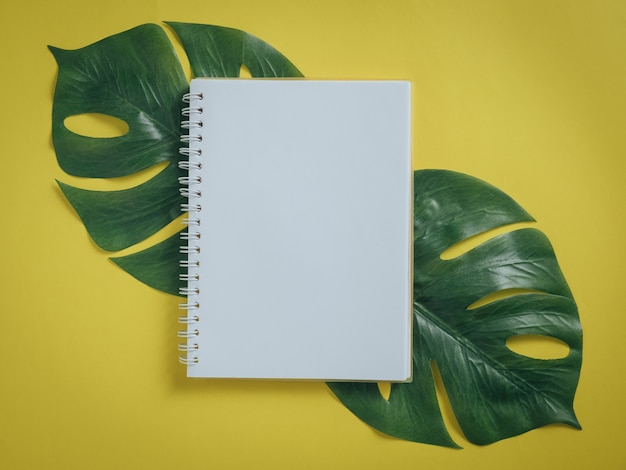 Escritorio de mesa de oficina con cuaderno, hojas tropicales Monstera sobre fondo amarillo.