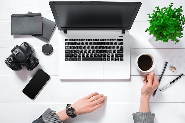 Escritorio de mesa de oficina con vista superior y endecha plana. Espacio de trabajo con manos de niña, portátil, flor verde en una olla, diario negro, taza de café sobre fondo blanco.