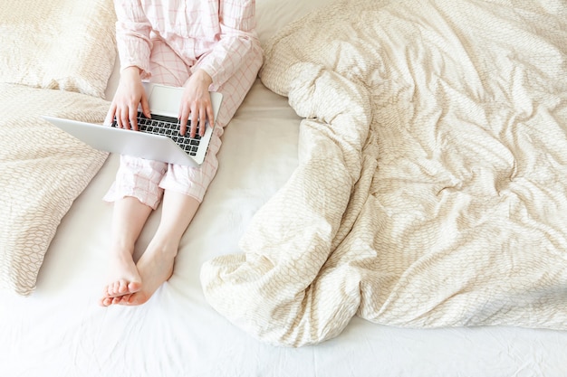 Escritório móvel em casa. Mulher nova nos pijamas que sentam-se na cama em casa que trabalha usando-se no computador do PC portátil. Garota de estilo de vida estudando dentro de casa. Conceito de quarentena de negócios freelance.