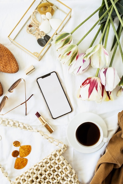 Escritorio de oficina en casa de blogger de belleza. Teléfono inteligente de pantalla en blanco, ramo de flores de tulipán, ropa y accesorios en lino blanco. Endecha plana