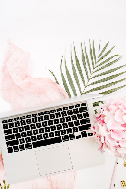 Escritorio de oficina en casa femenino con computadora portátil, ramo de flores de hortensia rosa, manta pastel, placa de hoja de monstera y accesorios en superficie blanca