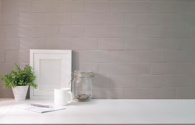 Escritorio de oficina en casa con marco de fotos, cuaderno de taza de café y planta de interior en mesa blanca con luz solar a través de persianas junto a la ventana