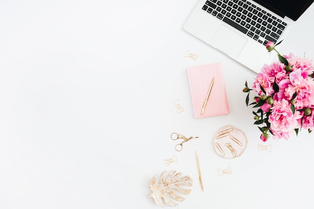 Escritorio de oficina en casa plano. Espacio de trabajo de mujer con laptop, ramo de peonías rosas, accesorios dorados, diario rosa sobre blanco