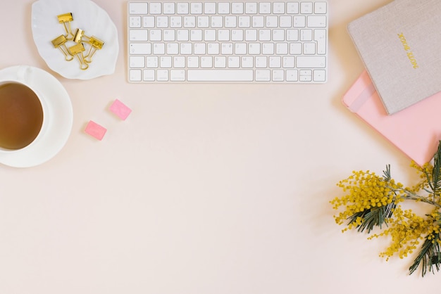 Escritorio de oficina cuadernos teclado y lápices de colores una taza de té flores de mimosa sobre un fondo beige Concepto de negocio mínimo para mujeres Espacio de copia de vista superior plana