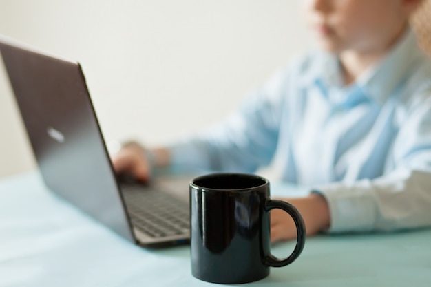 Escritorio de oficina de espacio de trabajo con laptop y taza de café. Pequeño muchacho lindo del negocio que se sienta en la tabla y que mira documentos importantes. Niño aprendiendo en línea.