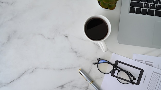 Escritorio de oficina con ordenador portátil, papeleo, vasos y taza de café sobre fondo de mármol.