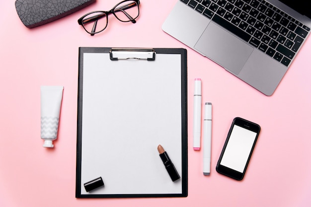 Escritorio de oficina rosa de mujer con hoja de papel limpia con espacio de copia libre, computadora portátil, teléfono con pantalla en blanco en blanco, crema, lápiz labial, anteojos y suministros de fondo.