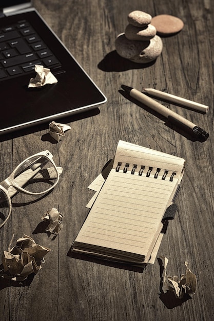 Escritura creativa, storyboard, bloqueo del escritor. Concepto de naturaleza muerta, arreglo sobre mesa de madera. Abra la libreta de papel en blanco. Páginas de papel arrugadas. Ordenador portátil, auriculares, gafas, pirámide de piedra, taza de café.