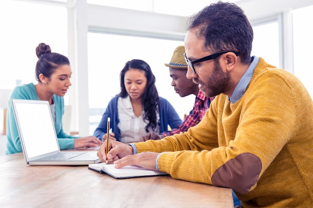 Escritura del hombre de negocios en libro mientras que se sienta con los colegas en oficina creativa