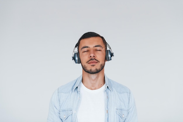 Escucha la música en los auriculares Hombre guapo joven de pie en el interior contra el fondo blanco.