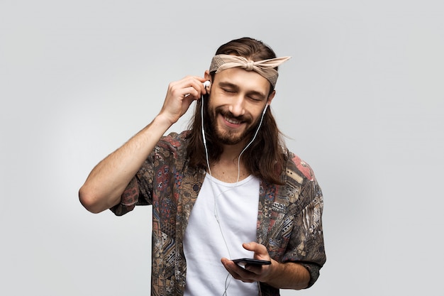 Escucha música y se relaja, disfruta, apuesto hombre hippie de pelo largo en auriculares con un teléfono inteligente en sus manos sobre un fondo blanco.