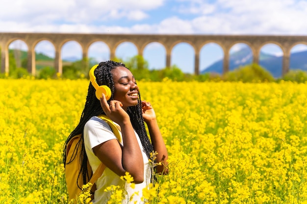 Escuchando música en auriculares amarillos con los ojos cerrados una chica étnica negra con trenzas un viajero en un campo de flores amarillas