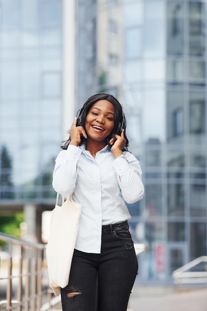 Escuchar música con auriculares Joven mujer afroamericana con camisa blanca al aire libre en la ciudad contra el edificio de negocios