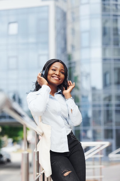 Escuchar música con auriculares Joven mujer afroamericana con camisa blanca al aire libre en la ciudad contra el edificio de negocios